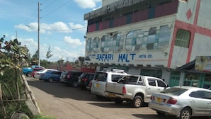 A parking lot with several vehicles, including a white pickup truck and a silver sedan, in front of a building with a sign that reads "SAFARI HALT HOTEL LTD." - Mt Kenya Safari Halt Hotel (Chaka)