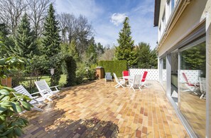 Un patio con piso de baldosas, sillones blancos y una puerta de cristal que da a una zona cubierta.