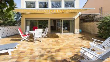 A patio area with a brick floor, white lounge chairs, a table with chairs, and a yellow awning.