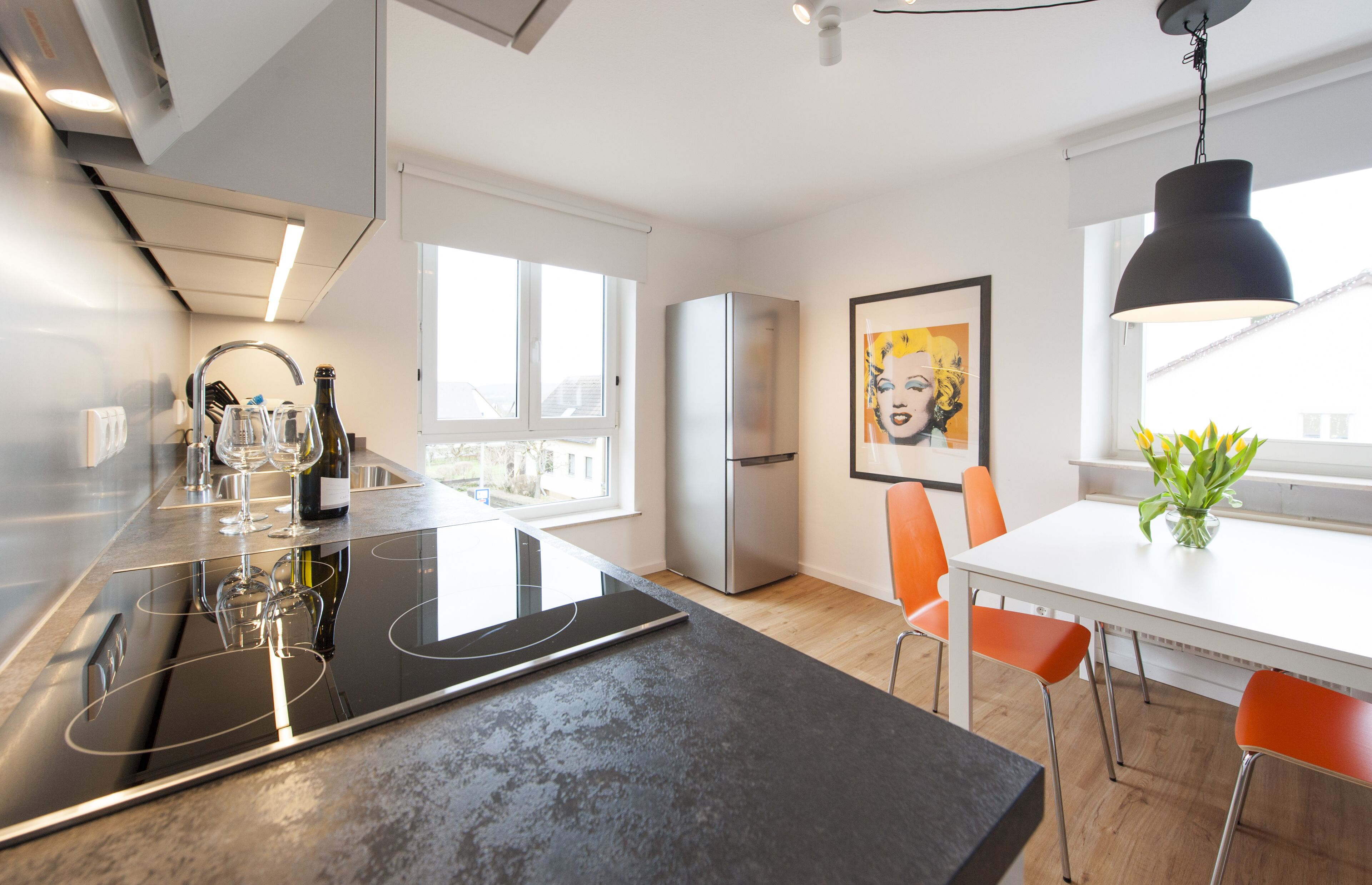 A modern kitchen with a built-in stove, a refrigerator, and a dining area with a white table and orange chairs.