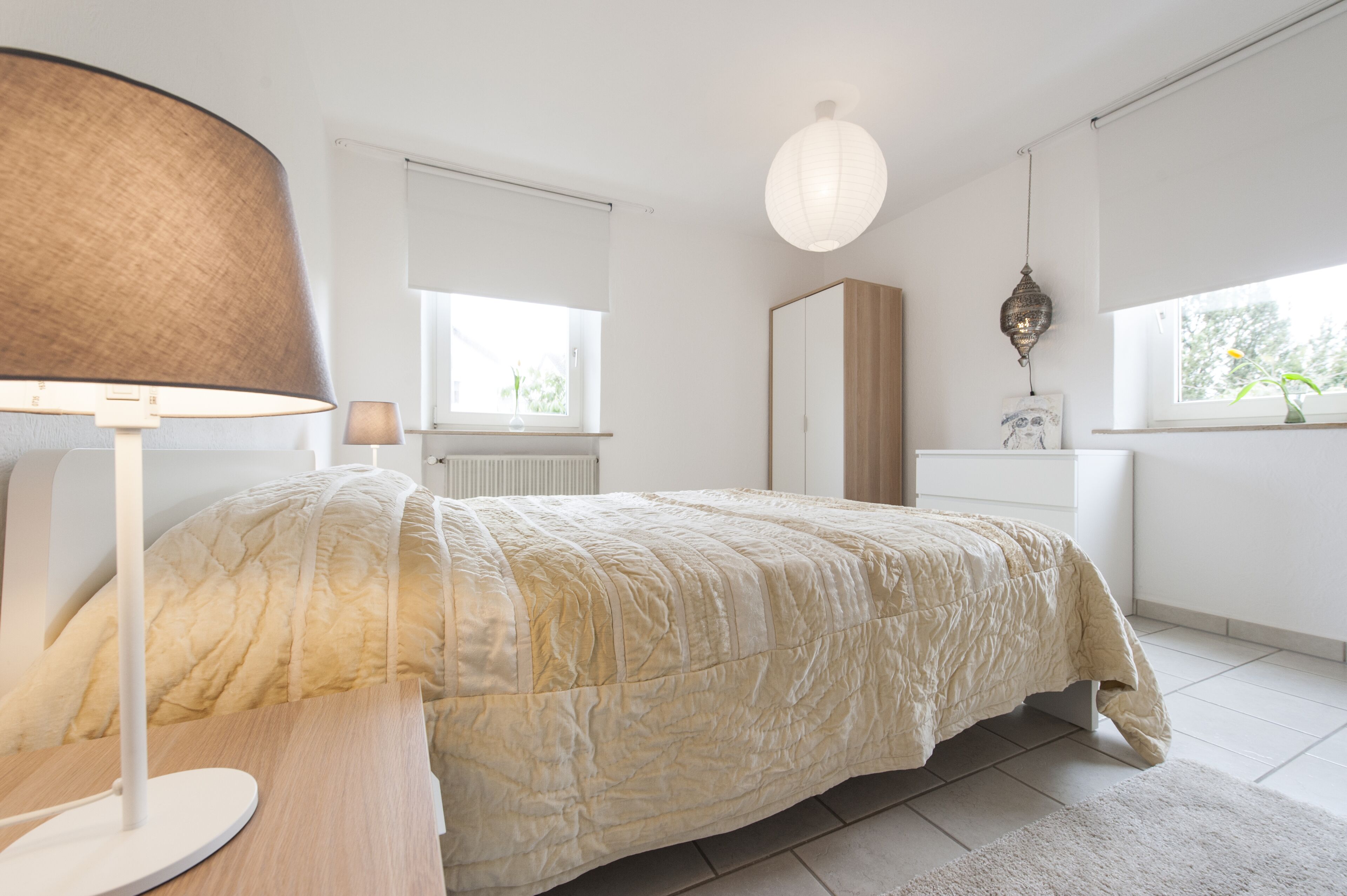 A bedroom with a bed, a lamp, a window, and a hanging light fixture.