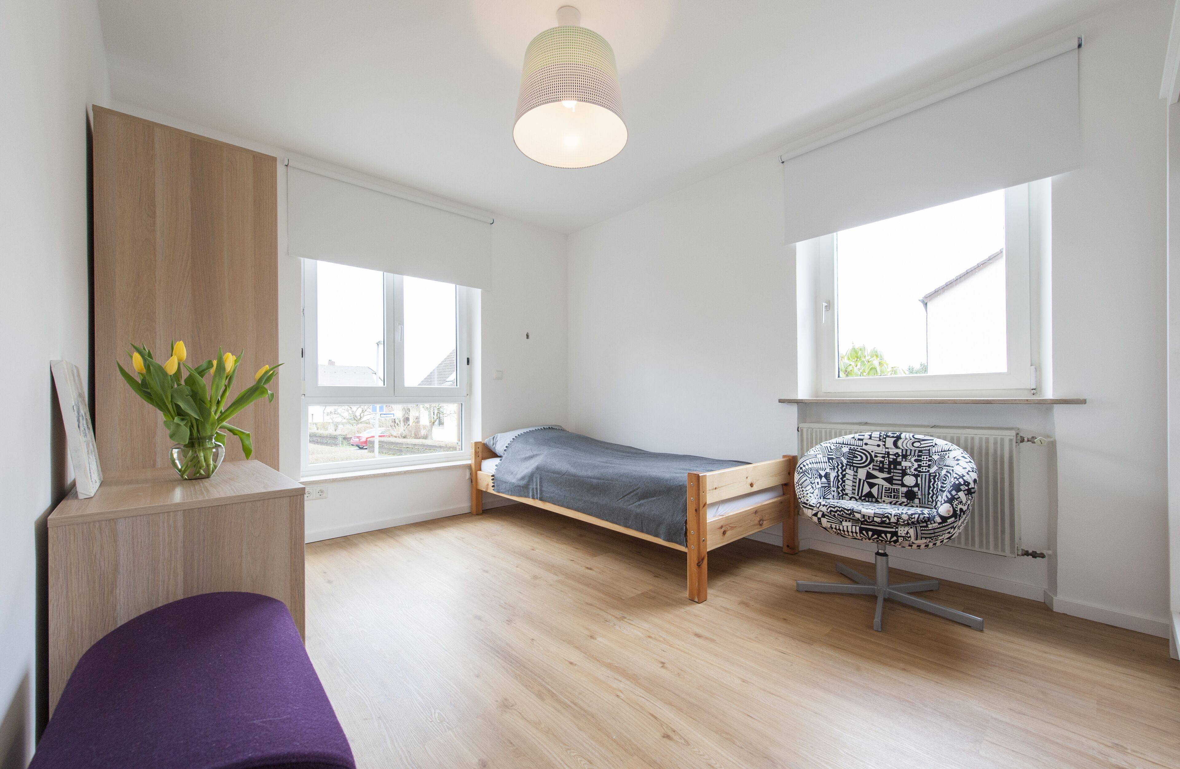 Quarto pequeno e luminoso com cama, escrivaninha e cadeira. Uma janela oferece luz natural e vista para o exterior.