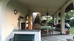 Uma varanda coberta com um banco verde, uma mesa de jantar com cadeiras, uma lareira e uma escadaria que dá para um andar superior.
