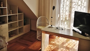 Quarto com teto inclinado, escrivaninha branca com monitor de computador, cadeira e microfone. Há uma estante com diversos objetos e uma janela com cortinas estampadas.