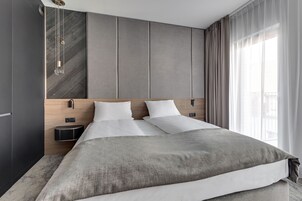A modern hotel room with a large bed, a wooden headboard, a bedside table, and a window with curtains.