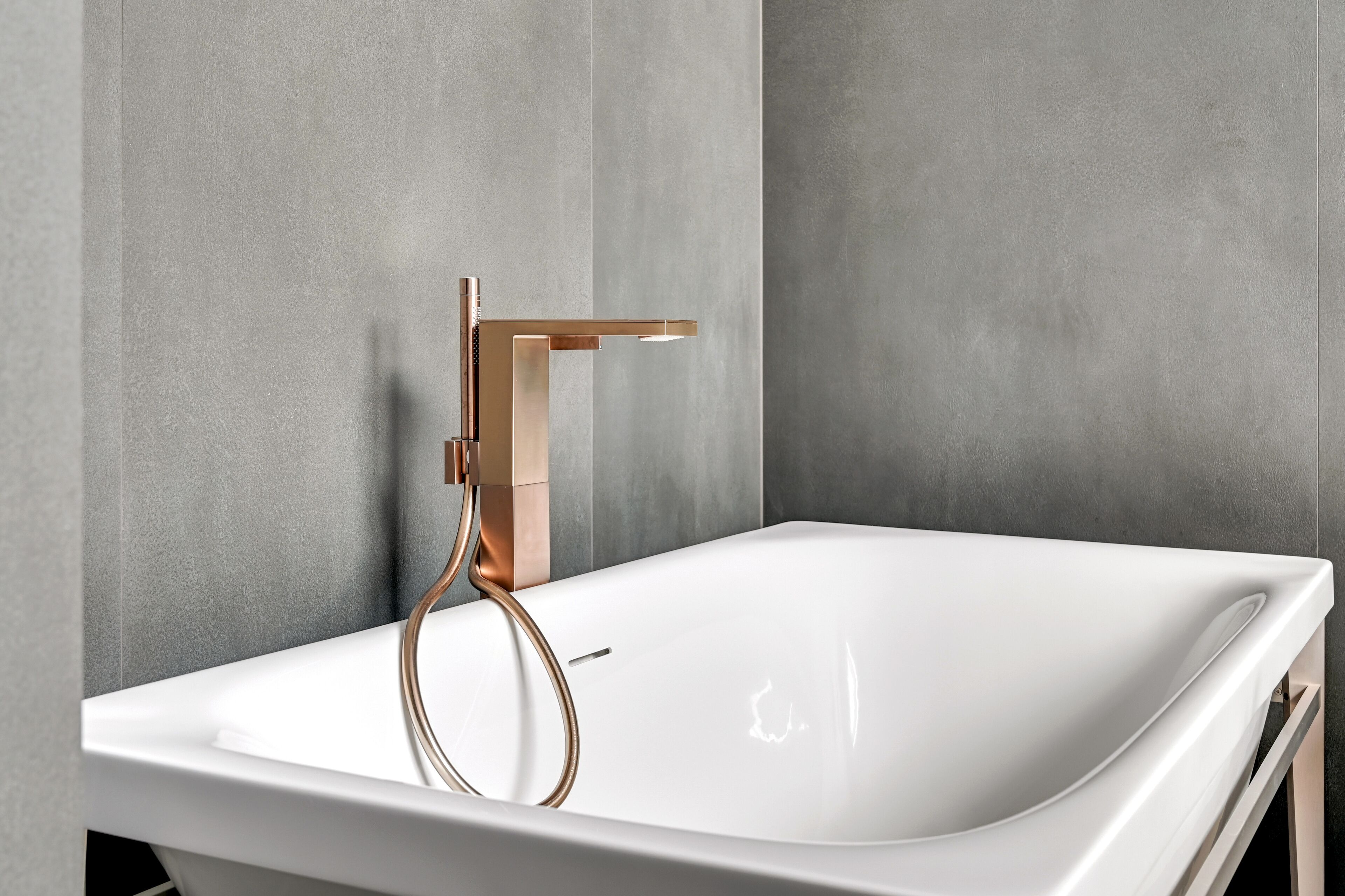 A modern bathroom with a white bathtub, copper faucet, and grey textured walls.