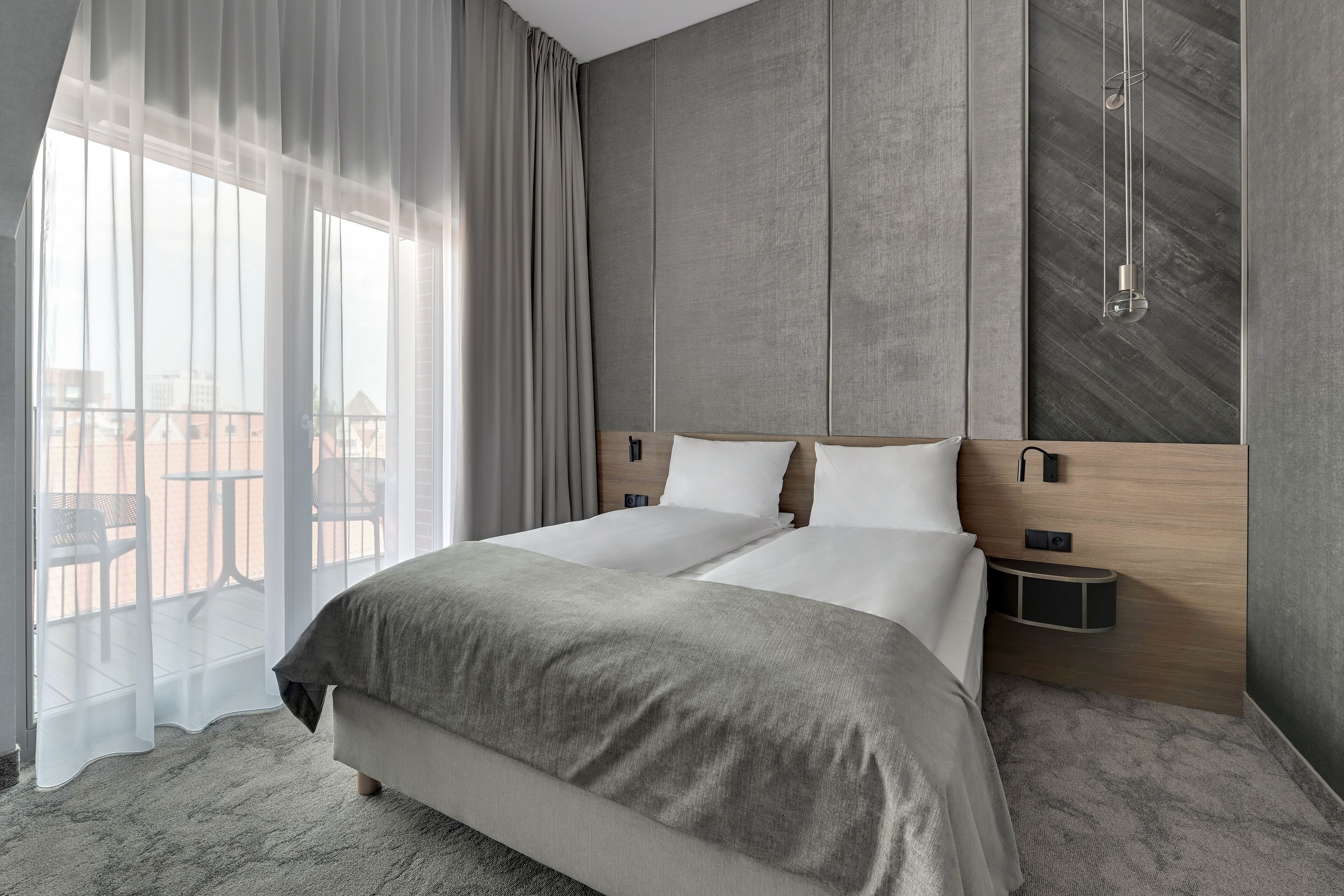 A modern hotel room with a large bed, a wooden headboard, a nightstand, and a view of a balcony.