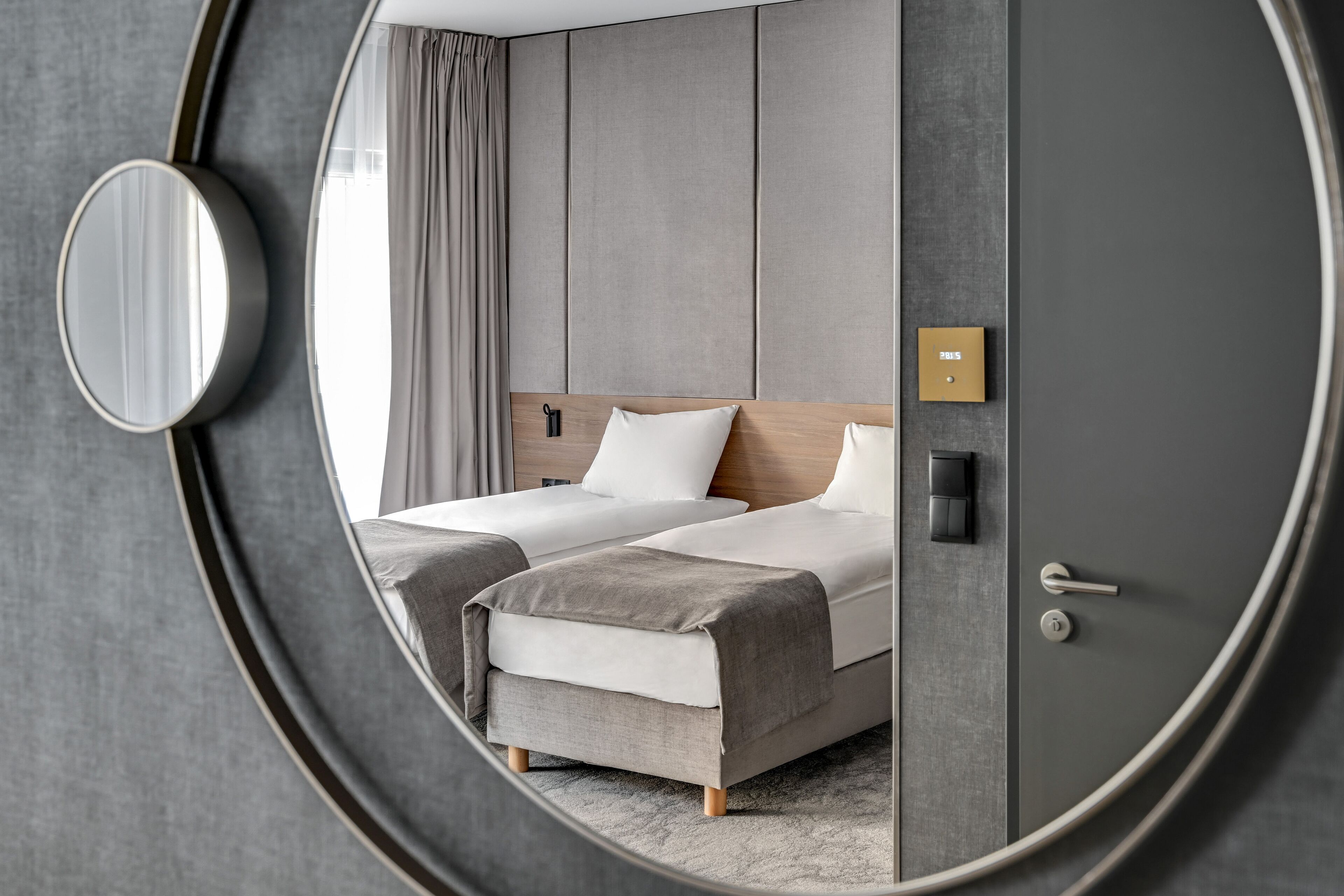 Una habitación de hotel moderna con un espejo redondo grande, dos camas con ropa de cama gris, un cabecero de madera, una mesita de noche y una puerta con pomo.