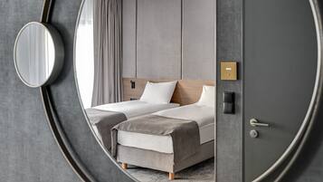 Una habitación de hotel moderna con un espejo redondo grande, dos camas con ropa de cama gris, un cabecero de madera, una mesita de noche y una puerta con pomo.