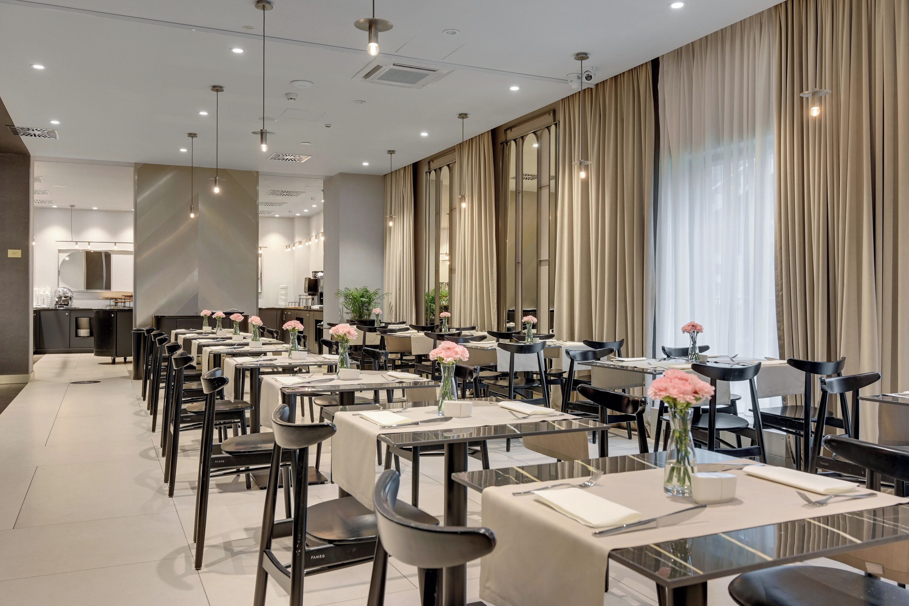 Un espace de restauration d’hôtel comprenant de nombreuses tables et chaises, de grandes fenêtres avec des rideaux et un aménagement intérieur moderne.