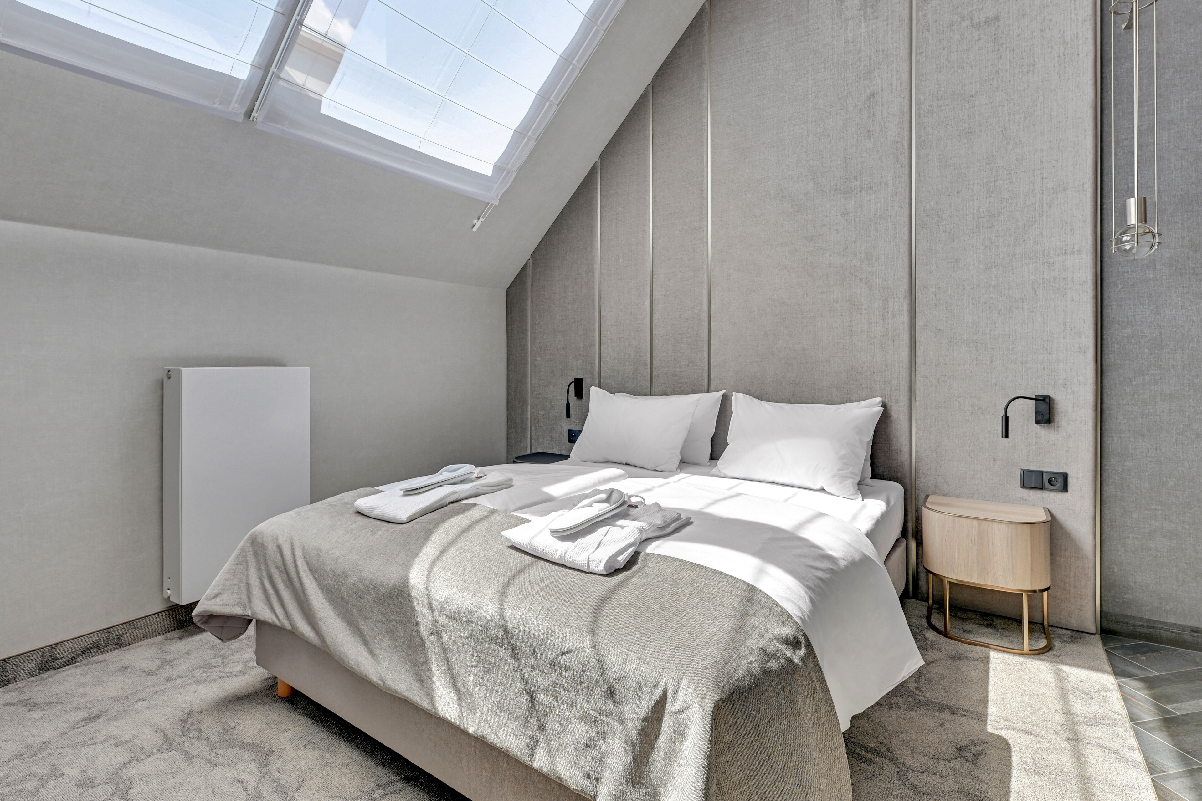 Une chambre moderne avec un puits de lumière, un lit sur lequel sont soigneusement pliées des serviettes, une table de chevet et un luminaire fixé au mur.