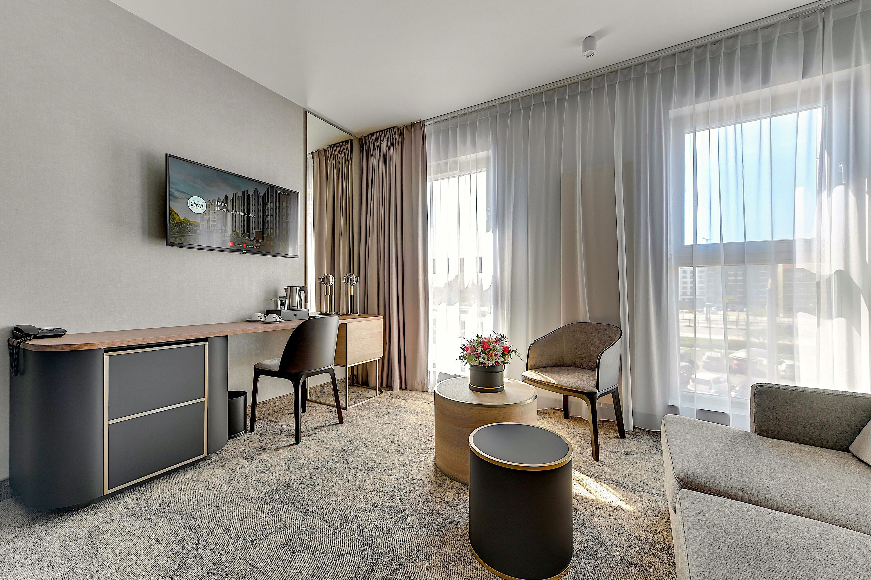 A modern hotel room with a desk, two chairs, a sofa, a round coffee table, and a large window with curtains.