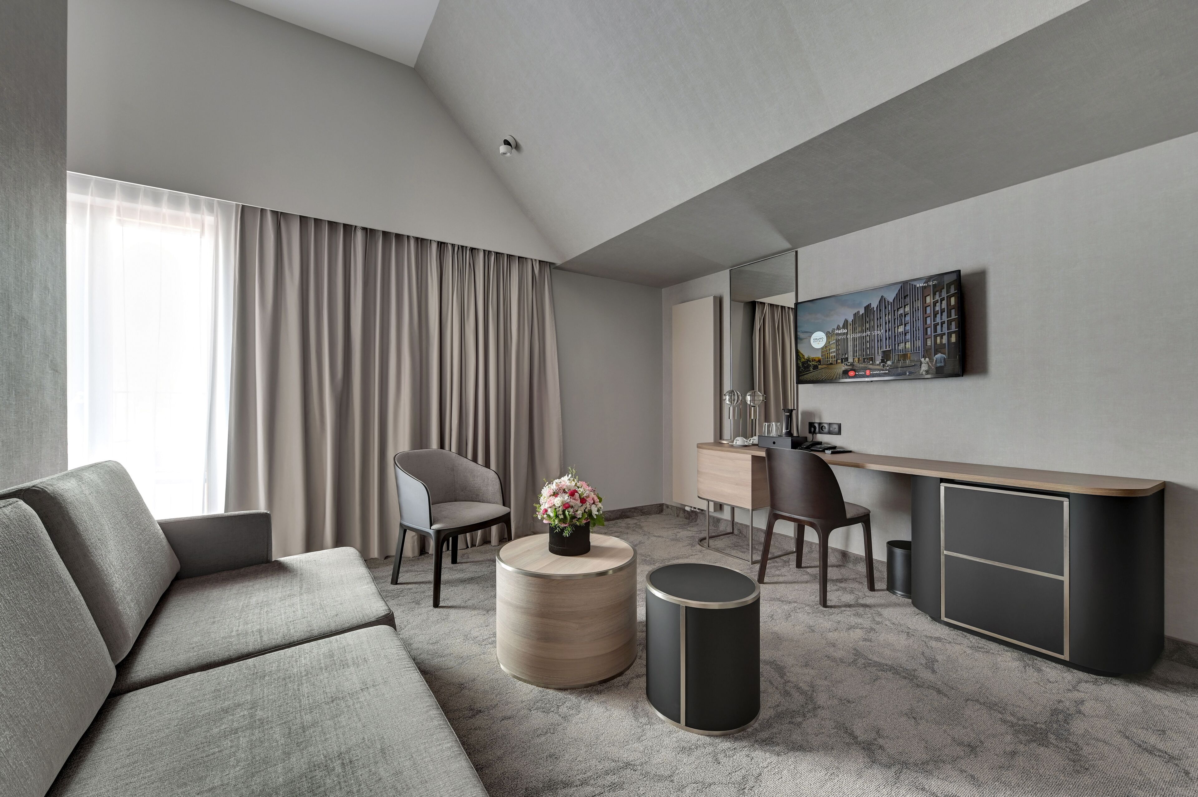 Ein modernes Hotelzimmer mit einem grauen Sofa, einem runden Holztisch mit Blumenvase, einem Schreibtisch mit Stuhl und einem großen Spiegel an der Wand.
