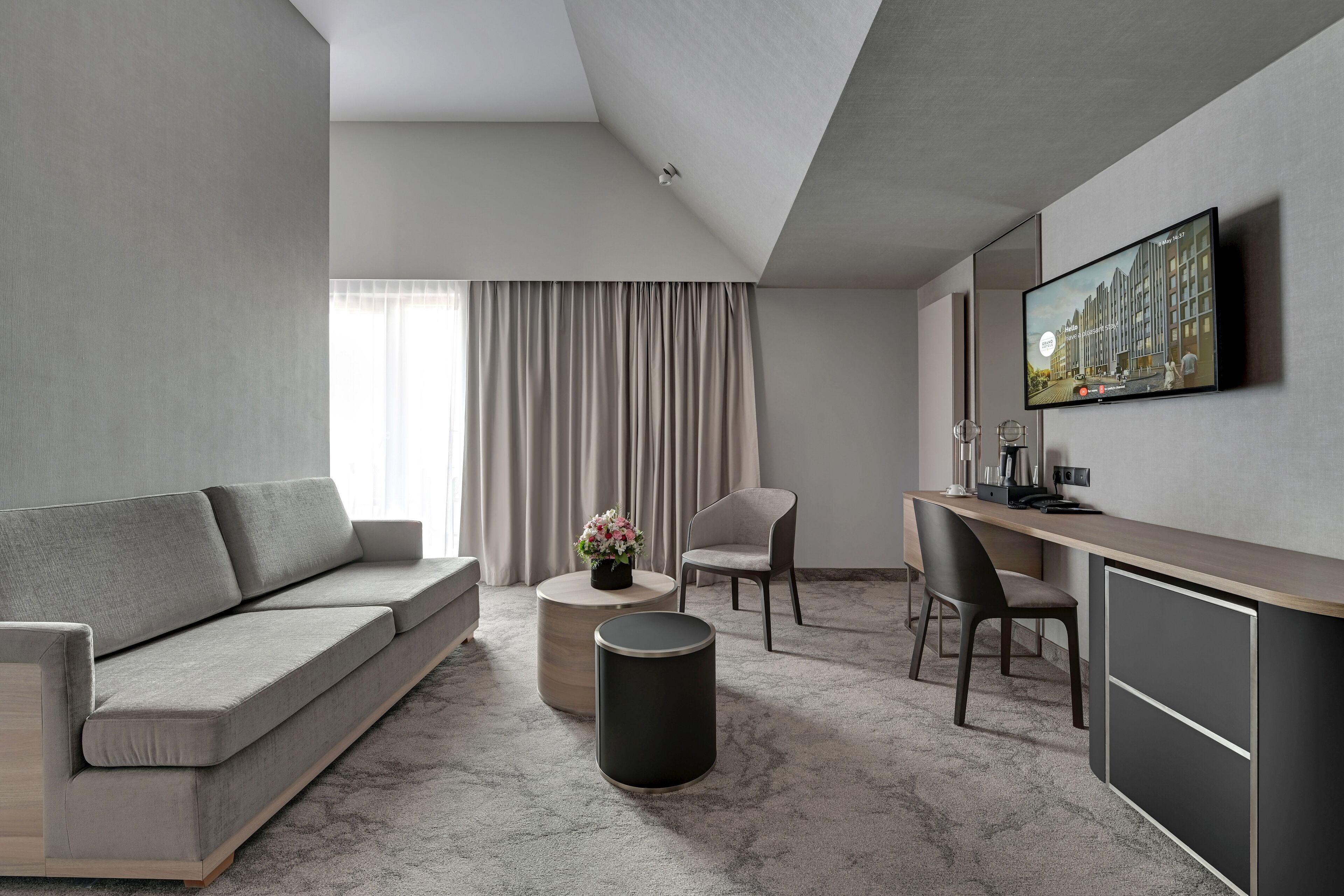 A modern hotel room with a sofa, a round coffee table with a vase, a desk with a chair, and a television mounted on the wall.