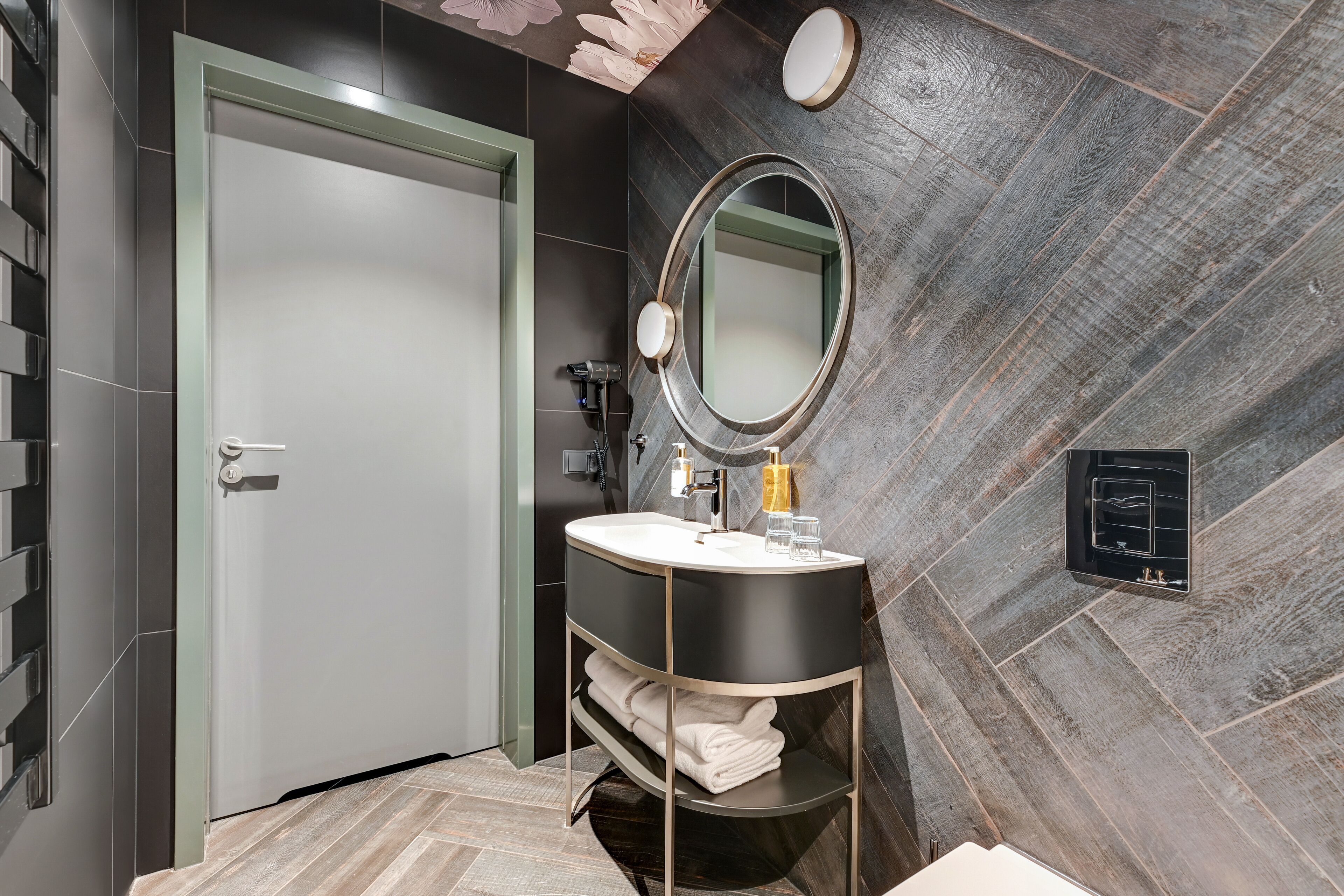 A modern bathroom with a grey door, a round mirror, a black vanity, and a herringbone-patterned wall.