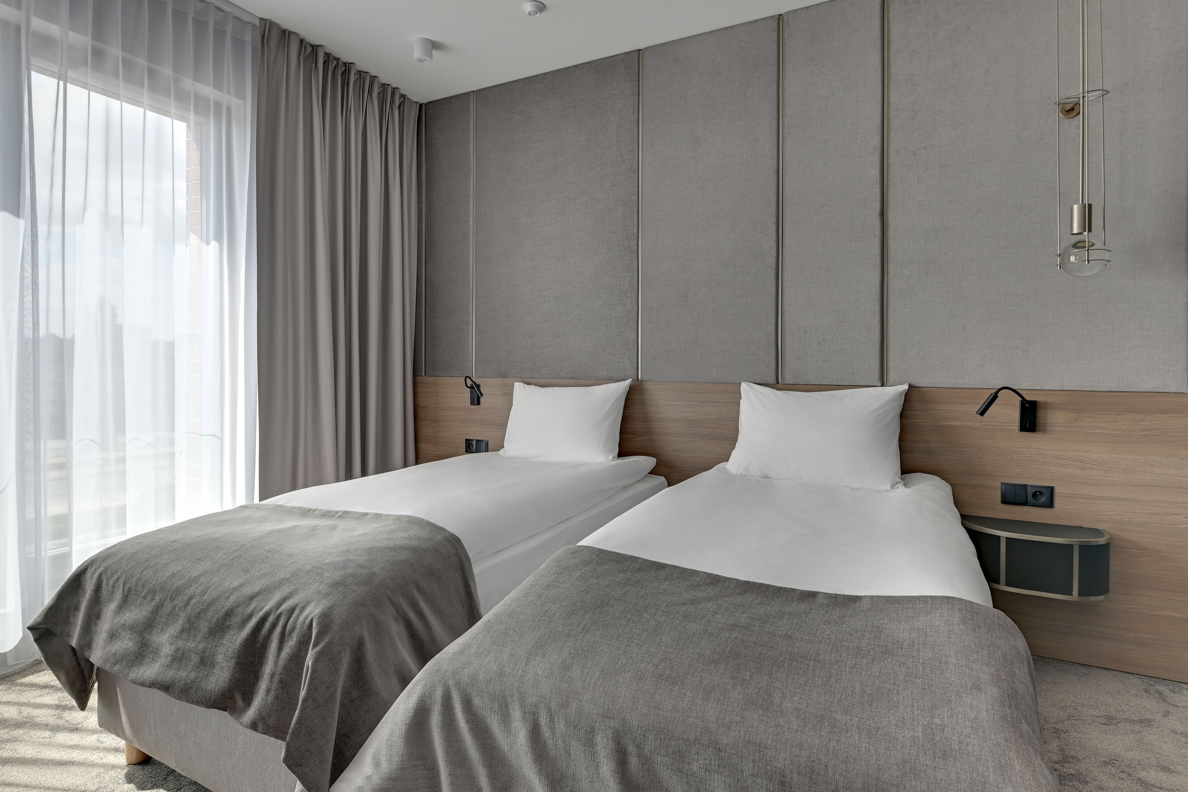A modern hotel room with two beds, a wooden headboard, a nightstand, and a large window with sheer curtains.