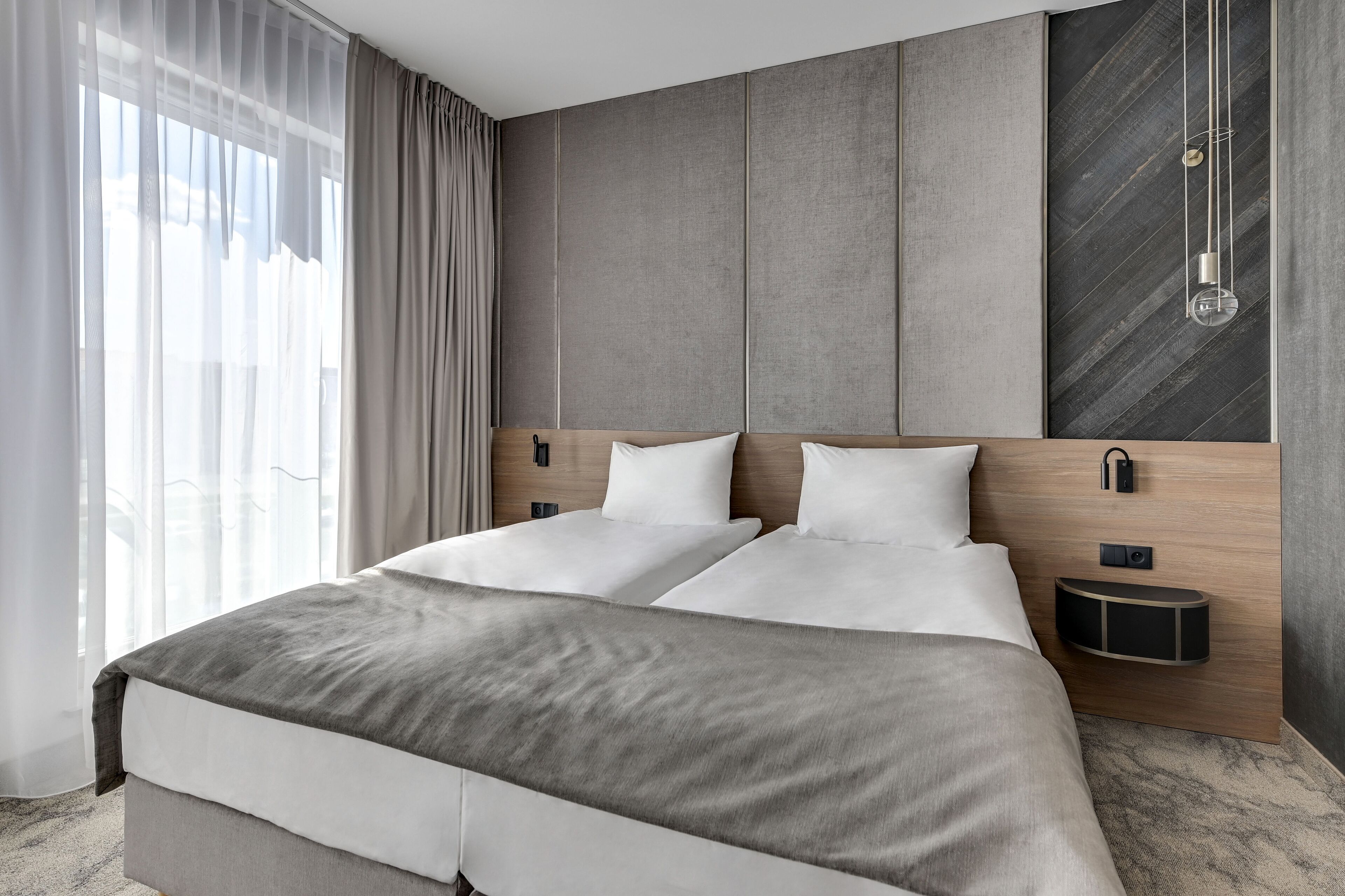 A modern hotel room with a large bed, a wooden headboard, a bedside table, and a wall-mounted light fixture.