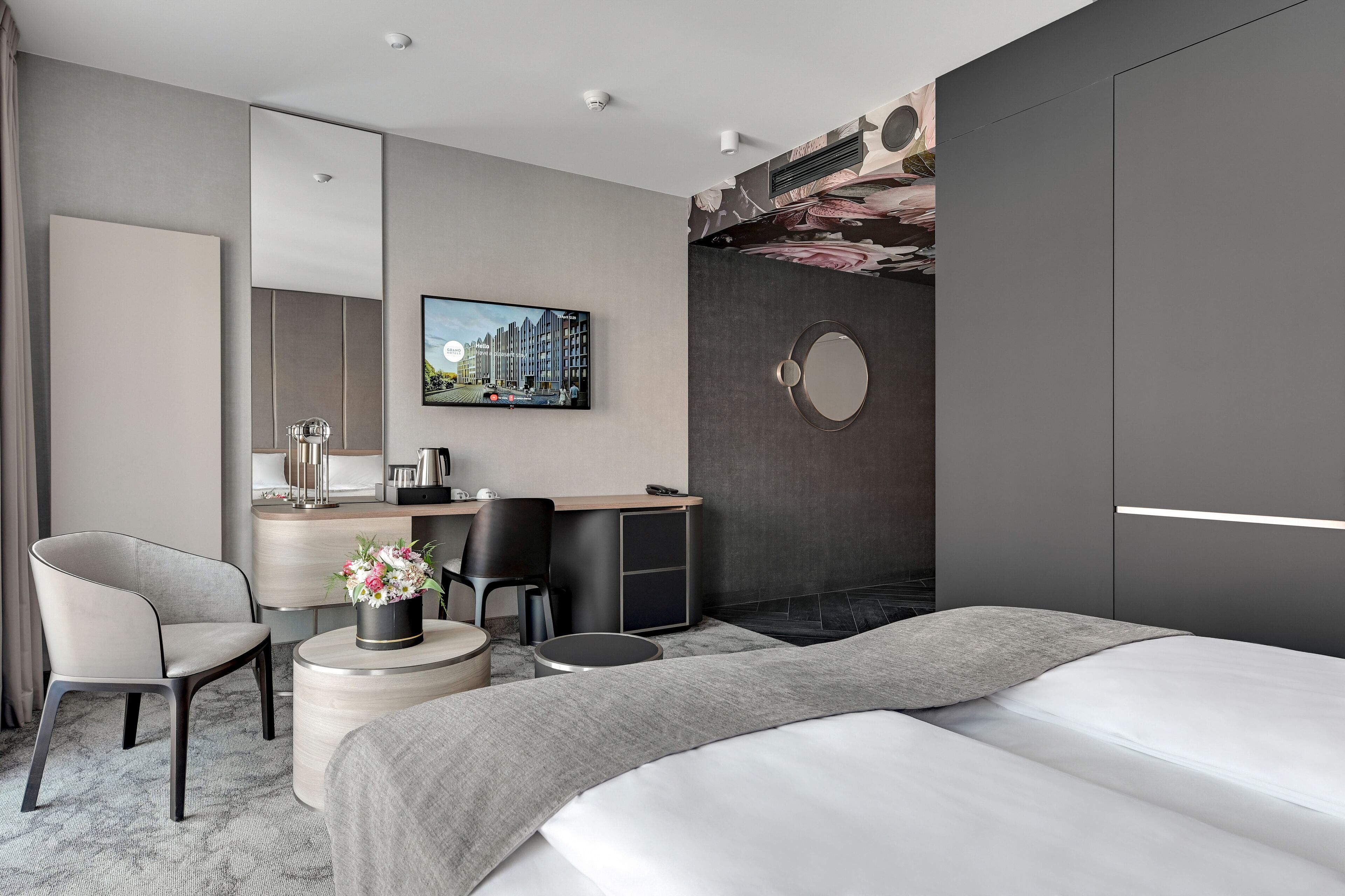 A modern hotel room with a large bed, a desk with a chair, a round mirror, and a television on the wall.