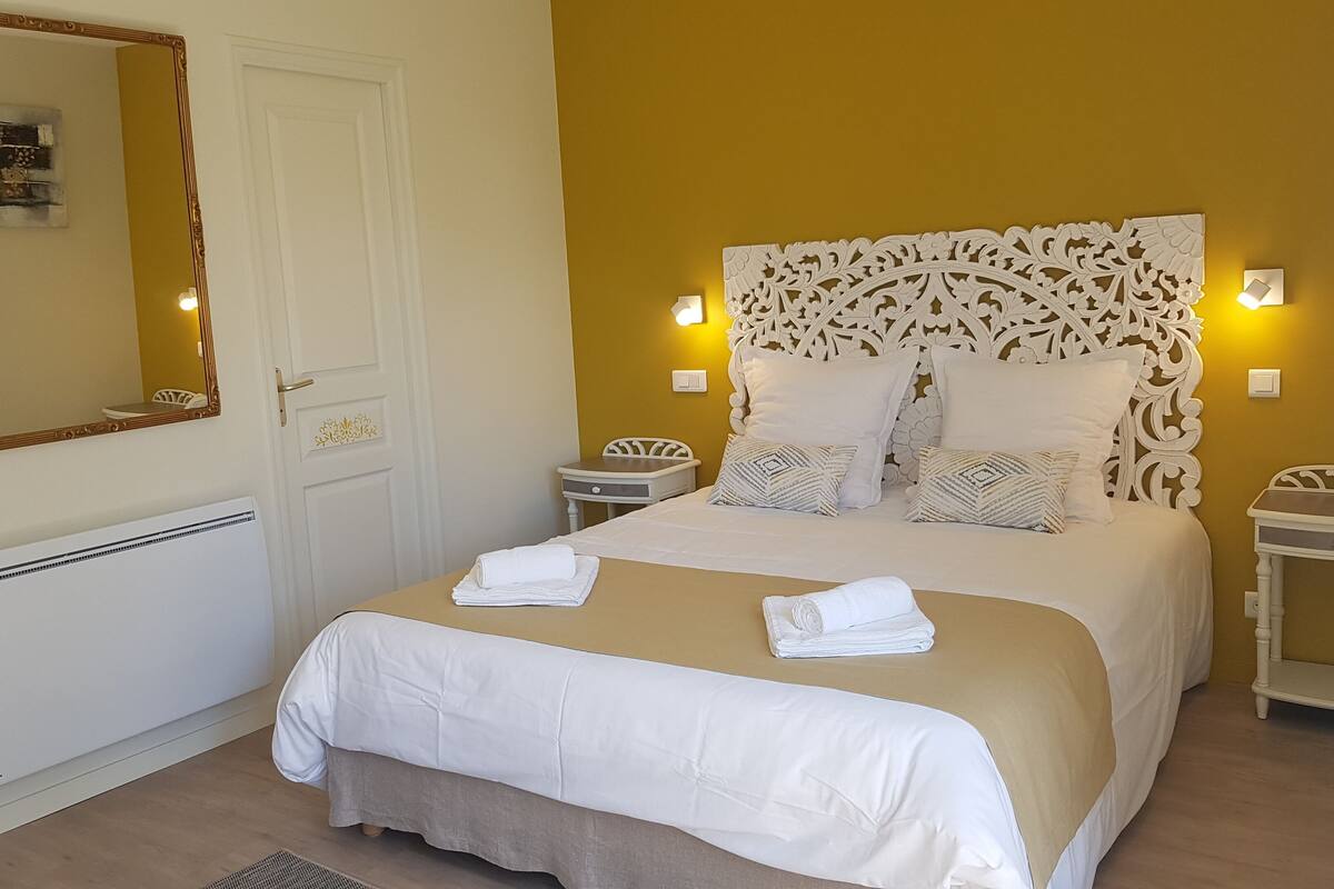 Een netjes opgemaakt bed met witte lakens en een beige sprei, met aan weerszijden twee nachtkastjes met lampen, een witte deur met decoratief paneel en een grote spiegel met gouden lijst.