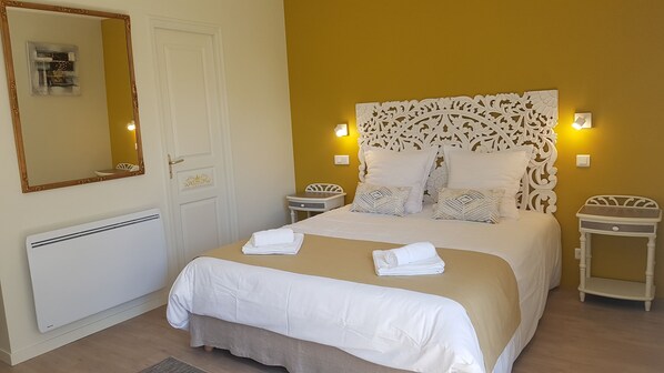 A neatly made bed with white linens and a beige bedspread, flanked by two bedside tables with lamps, a white door with a decorative panel, and a large mirror with a gold frame.