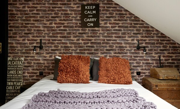 A bedroom with a brick wall, two brown pillows, a purple blanket, and a sign that says "KEEP CALM AND CARRY ON".