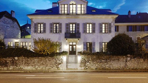 A large, two-story building with a symmetrical facade, a central entrance with a decorative door, and a stone wall in front. - Le Boutik Hotel (Annecy)