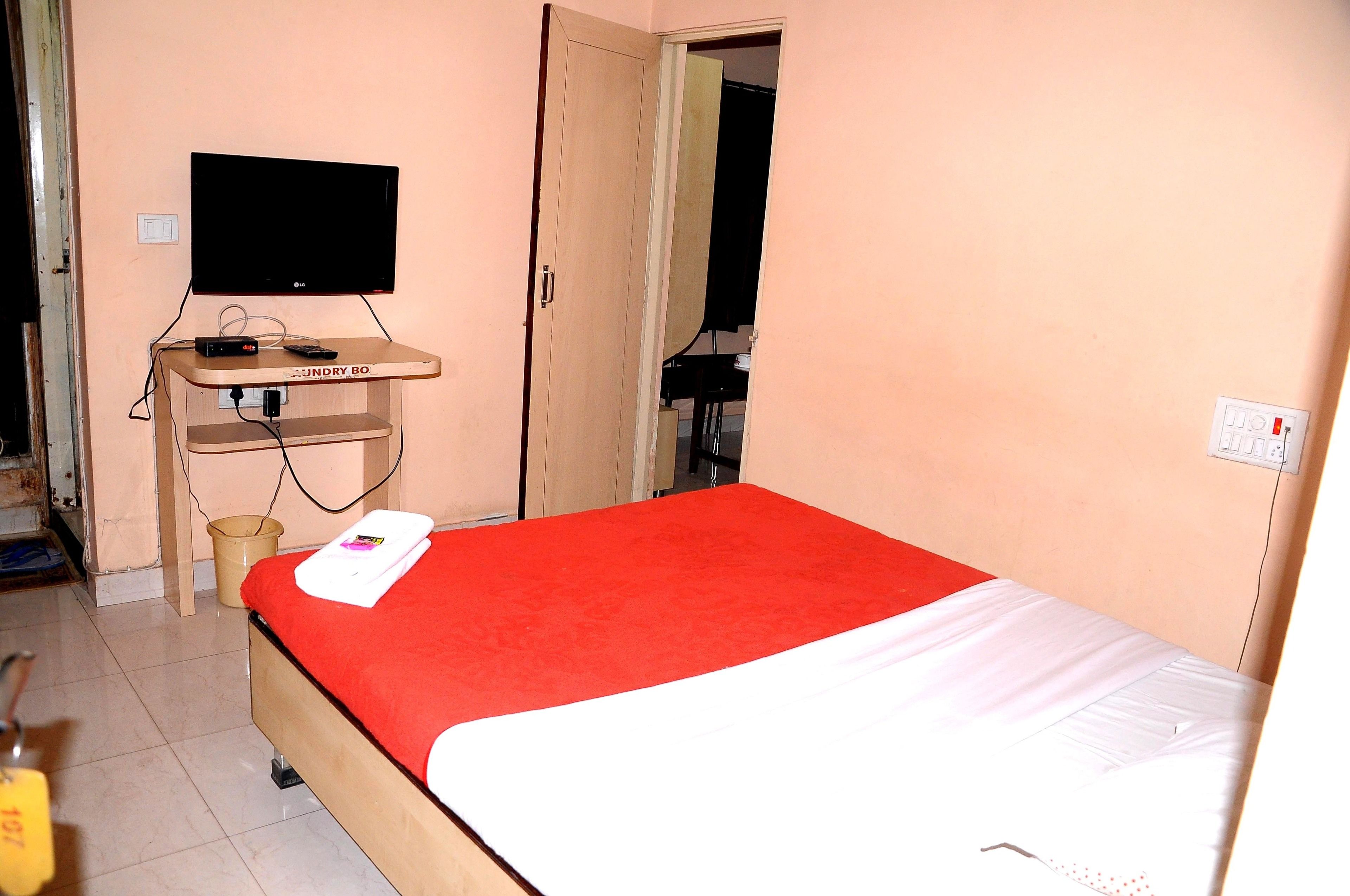 A bedroom with a bed, a TV on a stand, a small table, and a door leading to another room.