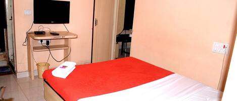 A bedroom with a bed, a TV on a stand, a small table, and a door leading to another room.