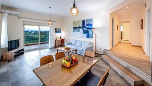 A modern living room with a dining area, a sofa, a TV, and a balcony with a city view. - Villa Alba with Private Pool & SeaView (Apolpena)