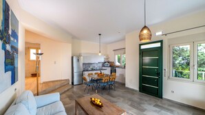 A modern living room with a sofa, a wooden coffee table with fruit, a dining area with chairs and a table, a kitchen with appliances, and a green door. - Villa Alba with Private Pool & SeaView (Apolpena)