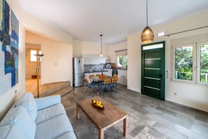 A modern living room with a sofa, a wooden coffee table with fruit, a dining area with chairs and a table, a kitchen with appliances, and a green door.