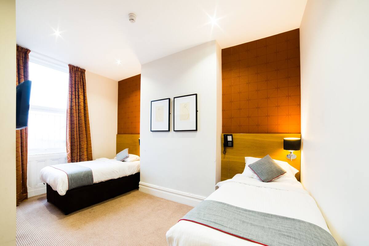 A hotel room with two beds, a wooden headboard, a wall-mounted phone, and a window with curtains.