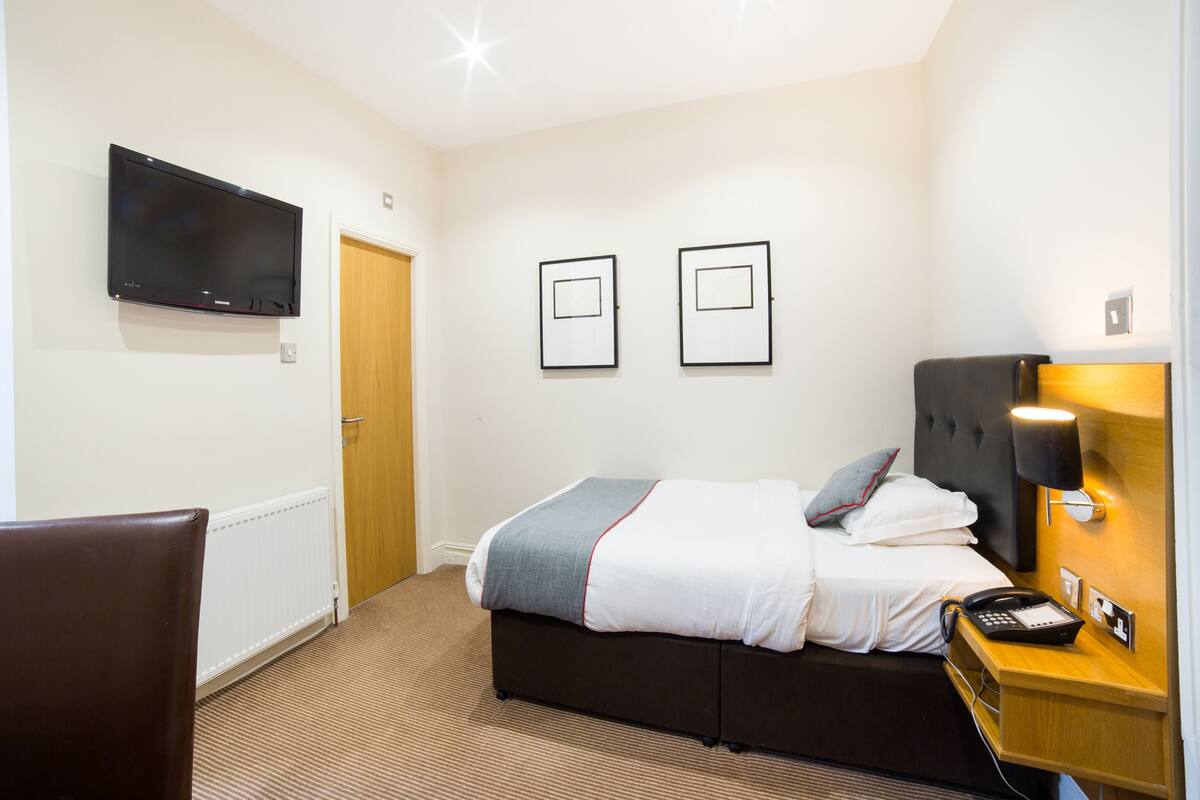 A hotel room with a bed, a TV mounted on the wall, a bedside table with a lamp, and a chair.
