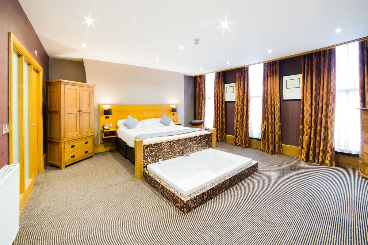 A hotel room with a large bed, a wooden wardrobe, a small desk, and a bathroom with a bathtub.