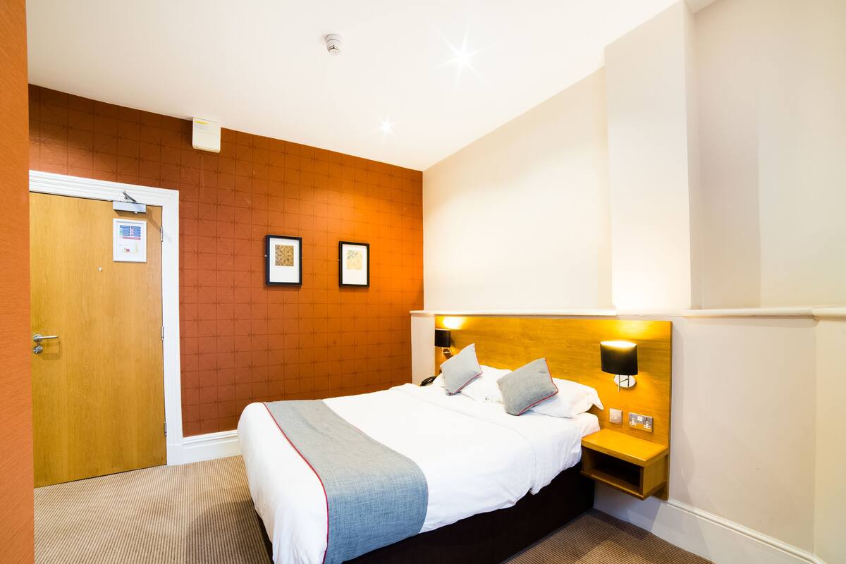 A hotel room with a bed, a wooden headboard, two bedside tables with lamps, a door, and a wall with two framed pictures.