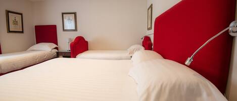 A hotel room with three single beds, each with white bedding and red headboards. There are framed pictures on the walls and a bedside lamp on the right side.