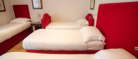 A hotel room with three beds, each with red headboards and white bedding. There are framed pictures on the walls and a small table with a lamp.