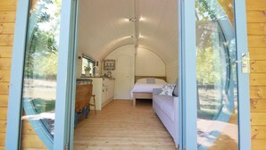 A cozy, compact room with a wooden floor, a bed with pillows, a small table, and a window. - Orchard Farm Luxury Glamping (Glastonbury)