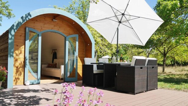 A wooden cabin with an open archway leading to a furnished outdoor area with a table and chairs. - Orchard Farm Luxury Glamping (Glastonbury)