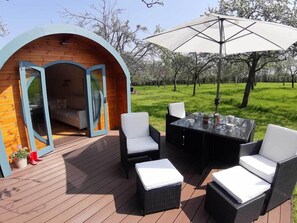 A cozy outdoor seating area with a wooden deck, a glass door entrance, and a large umbrella providing shade. - Orchard Farm Luxury Glamping (Glastonbury)