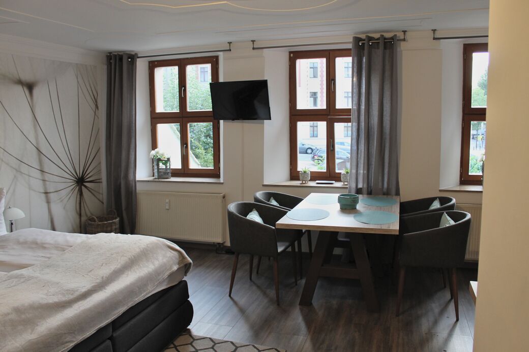 Una habitación de hotel con cama, una mesa de comedor con sillas, un televisor montado en la pared y ventanas grandes con cortinas.