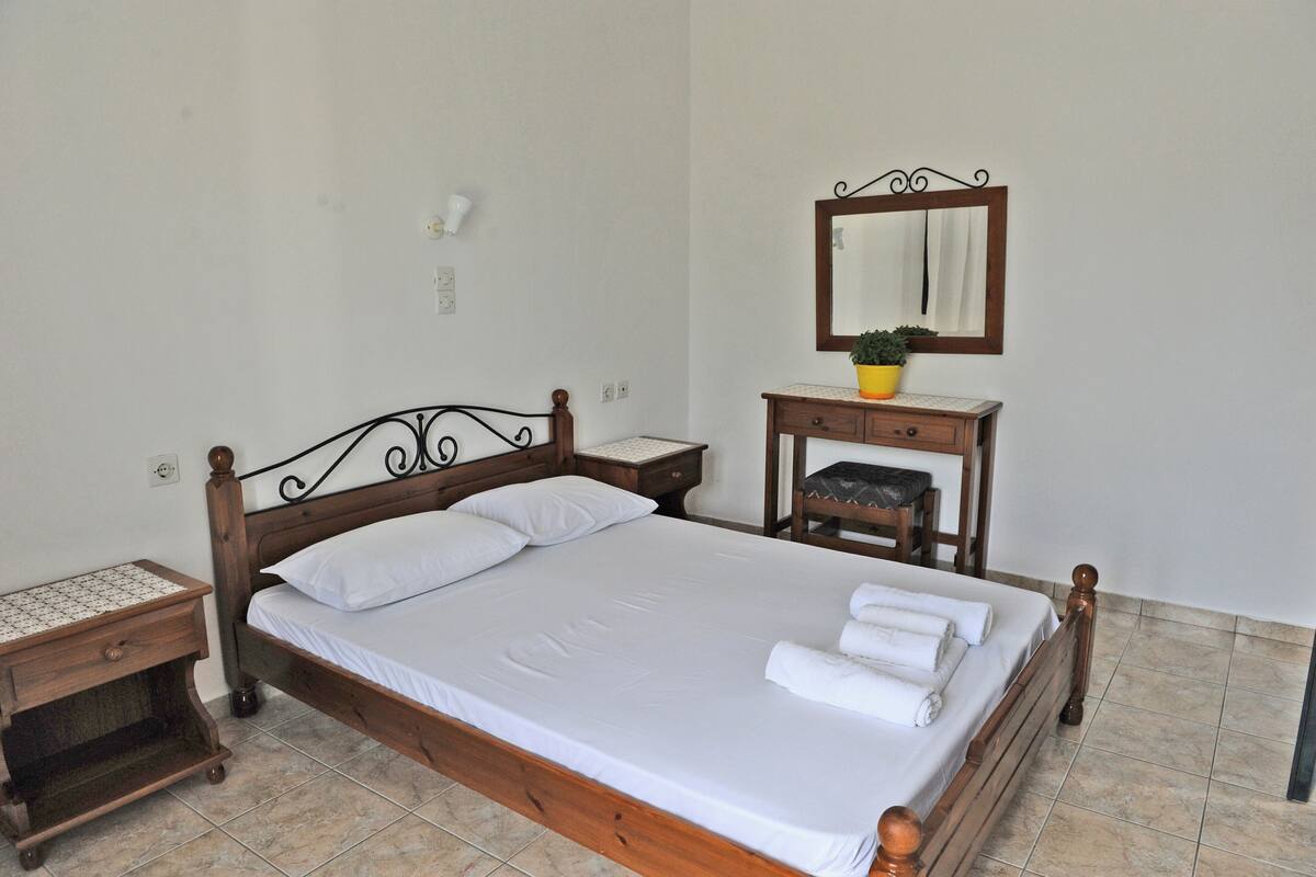 A neatly made bed with white linens and a wooden headboard, a bedside table with a lamp, and a small bedside table with a plant.