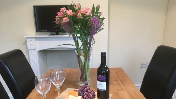 A dining table with a bottle of wine, two glasses, a cheese block, grapes, and a vase of flowers. - Old Shop (Newport)