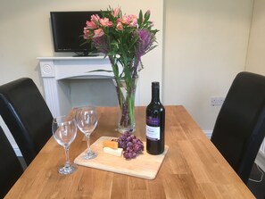 A dining table with a bottle of wine, two glasses, a cheese block, grapes, and a vase of flowers. - Old Shop (Newport)
