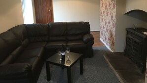 A living room with a black leather sofa, a small table with two wine glasses and a bottle, a wooden cabinet, and floral wallpaper. - Old Shop (Newport)