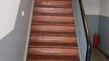 Un escalier en bois avec une main courante noire, des murs blancs et un extincteur rouge posé sur le sol.