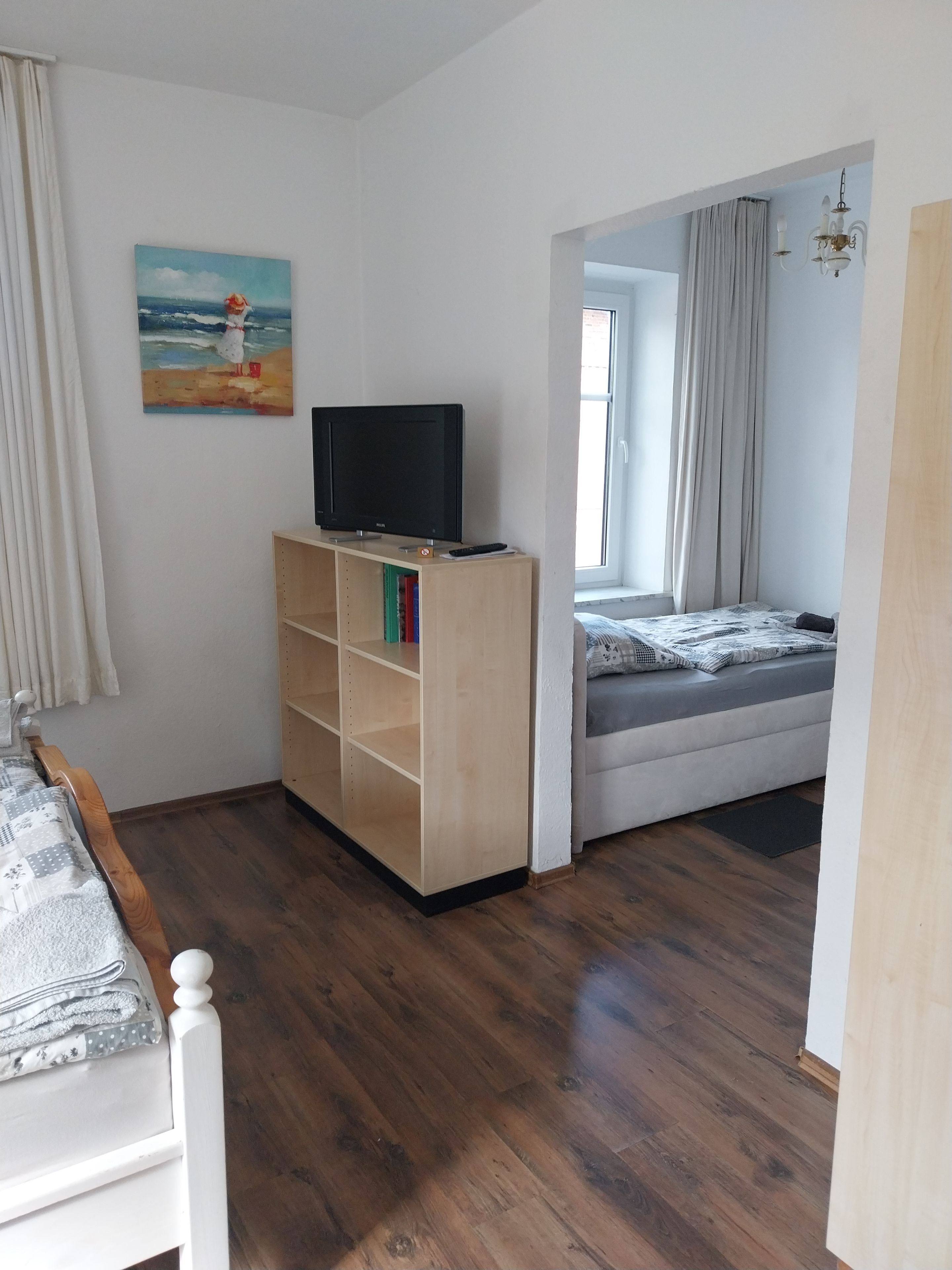 Ein Schlafzimmer mit einem Bett, einem Fernseher auf einem Holzschrank, einem Bild an der Wand und einem Fenster mit Vorhängen.