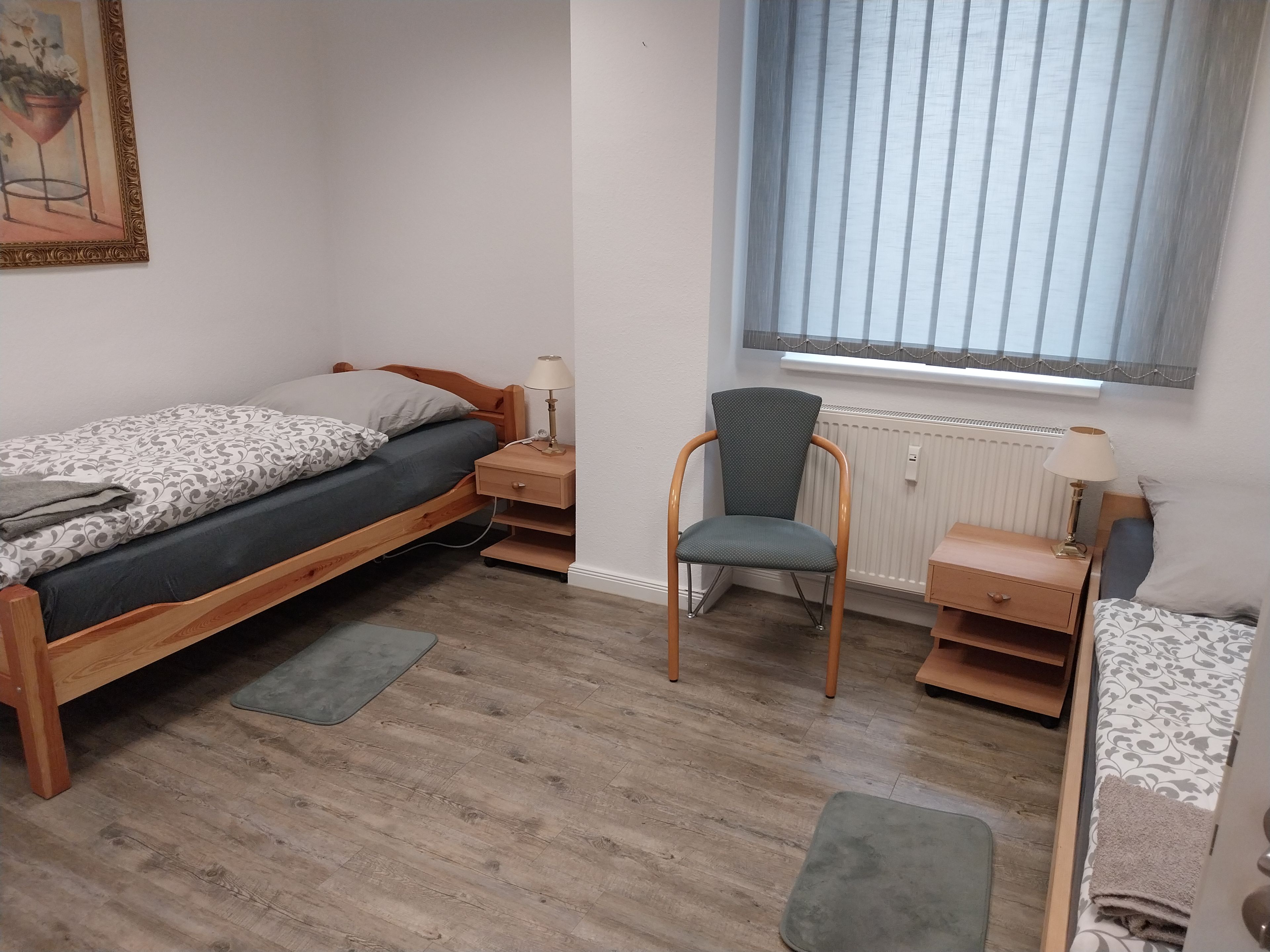 Quarto com cama, cadeira, mesa de cabeceira, janela com persianas e radiador.