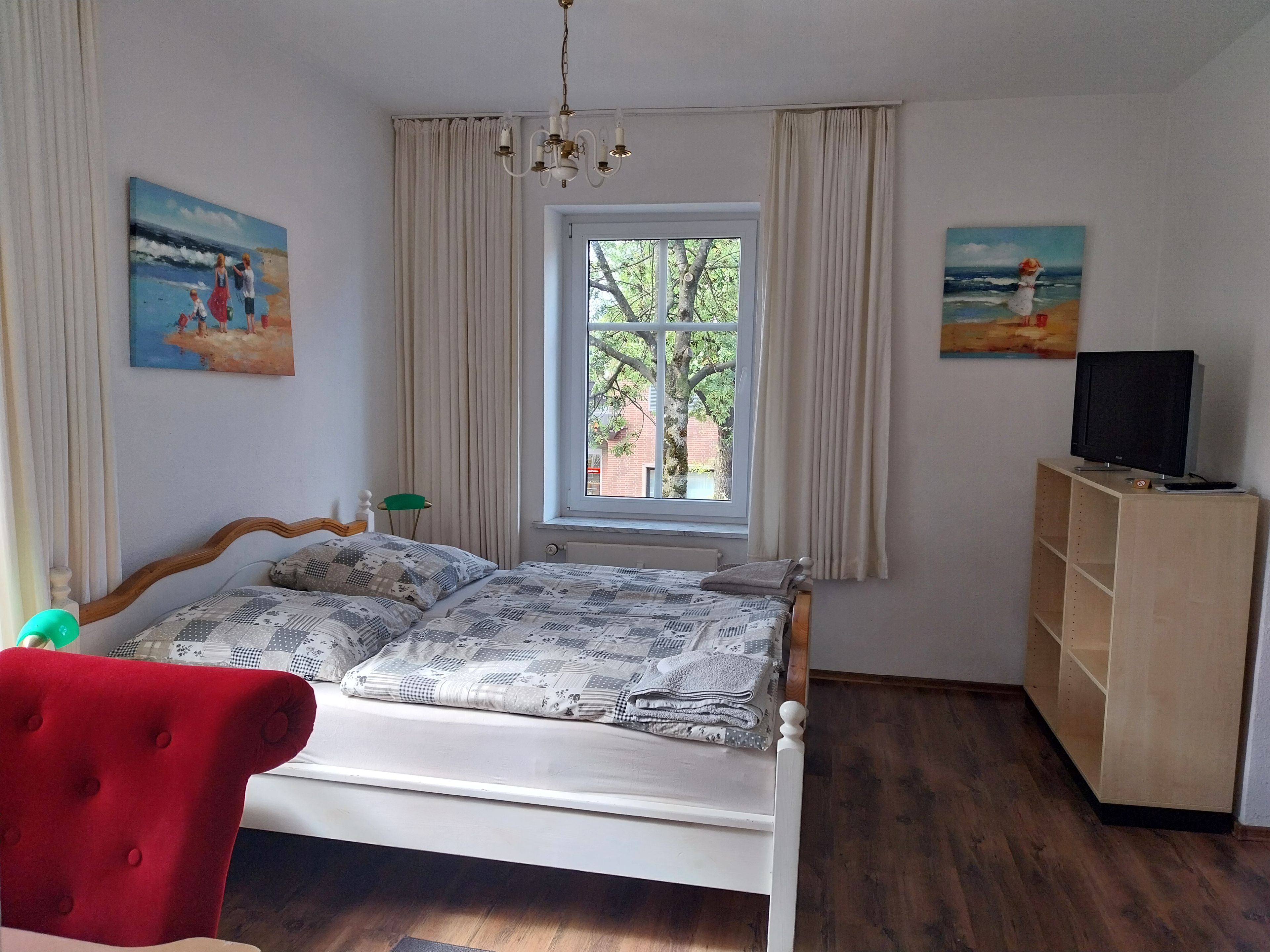 Un dormitorio con una cama, un televisor sobre un mueble bajo, una ventana con cortinas y dos cuadros en las paredes.