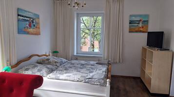 Un dormitorio con una cama, un televisor sobre un mueble bajo, una ventana con cortinas y dos cuadros en las paredes.